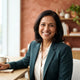A friendly South Asian woman smiling in a bright, professionally lit portrait with warm terracotta and burnt sienna backgroun